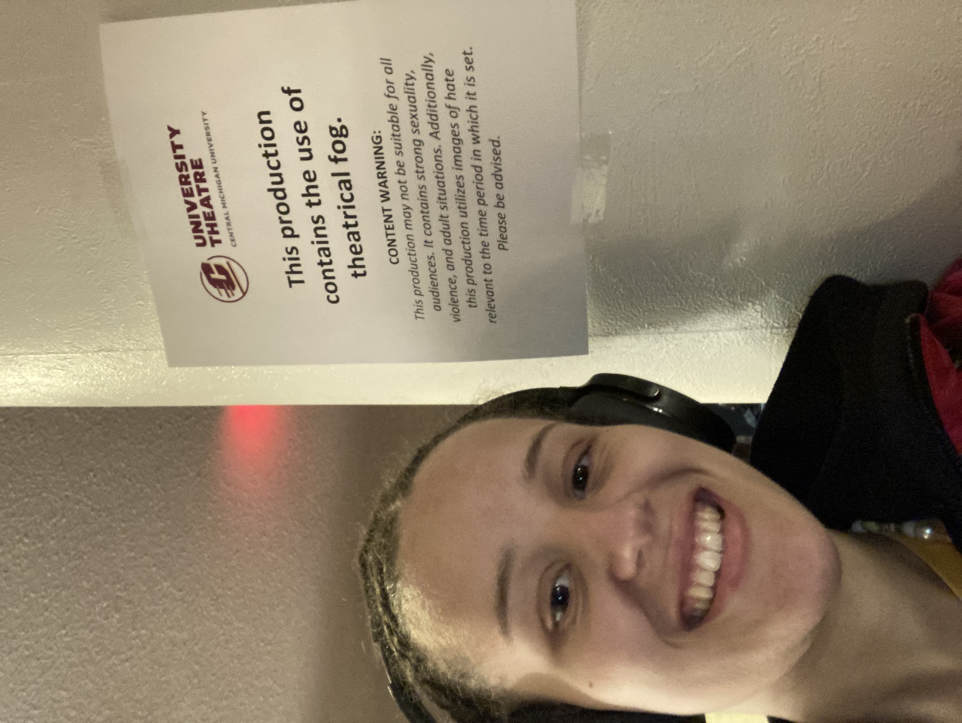 Picture of Gabrielle next to a posted piece of paper reading University Theater Central Michigan University. This Production contains the use of theatrical fog. Content warning this production may not be suitable for all audiences. It contains strong sexuality violence and adult situations. Additionally this production utilises images of hate relevant to the time in which it is set. Please be advised.'